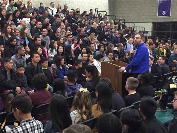 Luke Hackney speaks to a large group of parents and family members sitting on bleachers in a school gym.